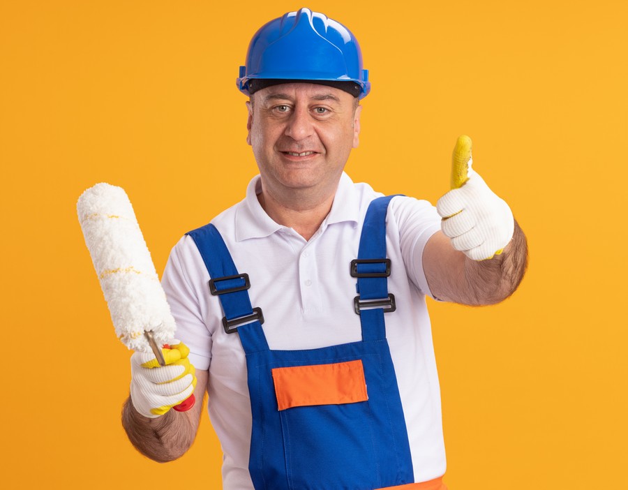 smiling caucasian adult builder man in uniform wearing protective gloves thumbs up and holds roller brush isolated on orange background with copy space